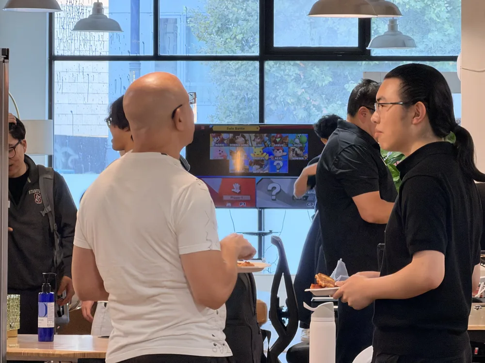 Attendees chatting over food in front of a TV showing the Super Smash Bros character select screen